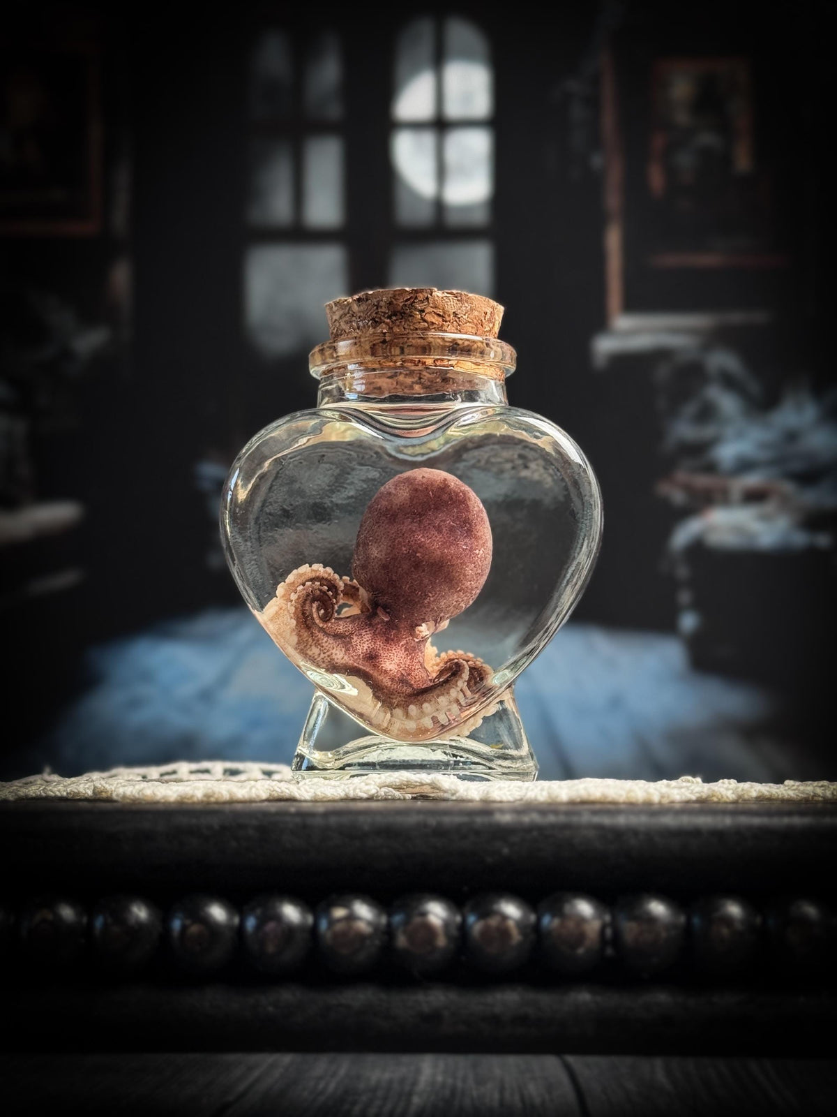 A small heart-shaped clear glass bottle with a cork stopper, containing a preserved octopus, displayed on a dark wooden surface against a moody, dimly lit room background.