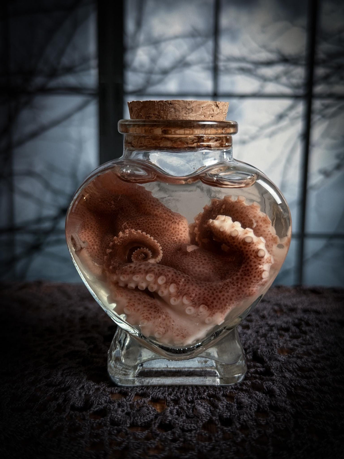 A preserved octopus inside a clear glass heart-shaped bottle with a cork stopper, set on a dark textured surface against a moody window background.