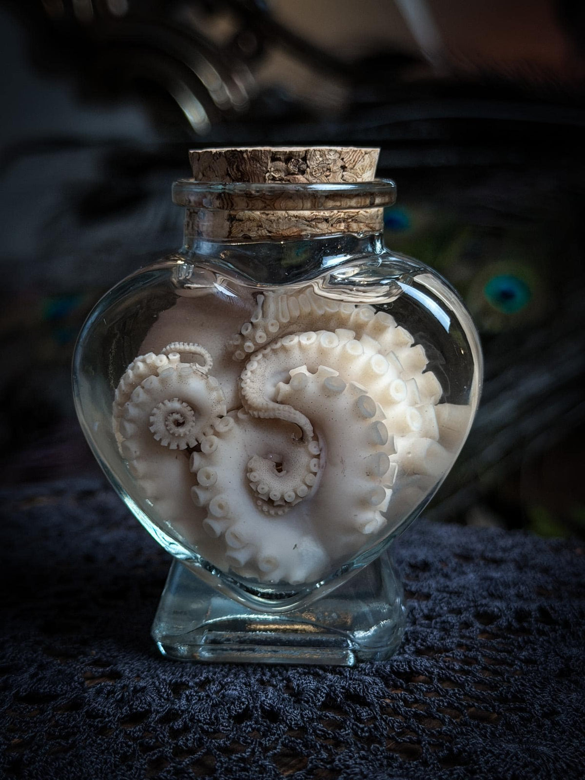 A heart-shaped clear glass bottle with a cork stopper, containing a white octopus-shaped object, set on a dark textured surface with a blurred dark background.