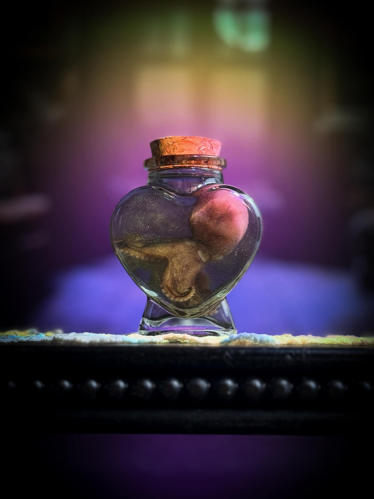A small heart-shaped clear glass vial with a cork stopper sits on a dark surface. Inside the vial is a preserved pinkish-brown specimen, photographed against a moody blurred purple and gold background.